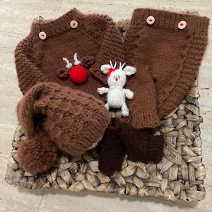 May include: A brown knitted baby outfit with a reindeer design, including a sweater, leggings, hat, and booties. The sweater has a red nose and white antlers. A small white reindeer toy is also included.