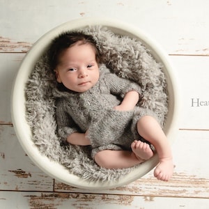 Könnte beinhalten: Ein neugeborenes Baby in einem grauen Strickoutfit liegt in einer weißen Schüssel, die mit einem grauen, flauschigen Material ausgekleidet ist. Das Baby schaut direkt in die Kamera.