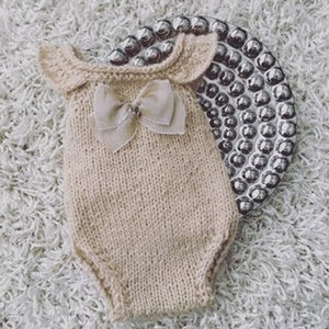May include: A beige knitted baby onesie with a bow and small wings. The onesie is made of a soft, comfortable material and is perfect for newborn photos.