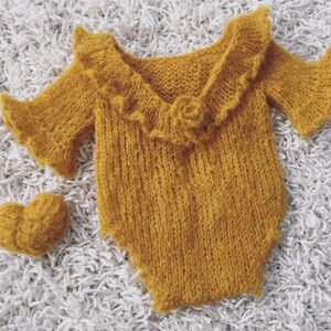 May include: A yellow knitted baby romper with a ruffled neckline and short sleeves. The romper is made of a soft, fuzzy yarn and has a heart-shaped knitted accessory.