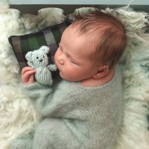 Könnte beinhalten: Ein Neugeborenes in einem hellblauen Strick-Outfit, das auf einer flauschigen, hellgrünen Decke liegt. Das Baby hält einen kleinen, hellblauen Strickteddybären. Ein kariertes Kissen stützt den Kopf des Babys. Das Bild enthält den Text "Obs Beauty Photography".
