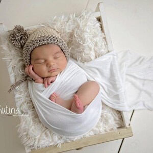 Op de afbeelding: Een pasgeboren baby slaapt rustig in een witte swaddle-deken. De baby draagt ​​een bruin en witte gebreide muts met een pompon bovenop. De baby ligt op een wit, pluche oppervlak in een houten doos.