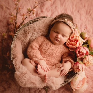 May include: A sleeping newborn baby wearing a pink knitted sweater and a headband, lying in a wicker basket lined with a pink fuzzy blanket. The baby is surrounded by pink peonies.