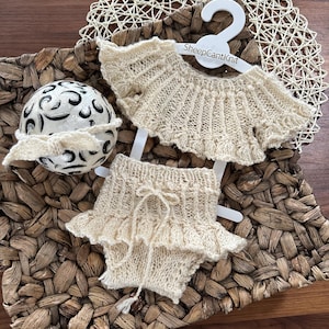 May include: Cream-colored knitted baby outfit, including a top and shorts, displayed on a white hanger. The top and shorts have a ruffled edge. The outfit is displayed on a woven basket. The text "SheepCantKnit" is on the hanger.