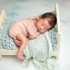 Op de afbeelding: Een pasgeboren baby slaapt op een wit houten bed met een lichtblauw gebreid dekentje. De baby draagt een roze gebreide romper.