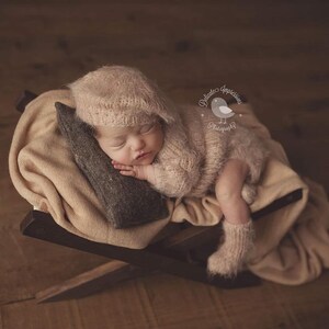 May include: A newborn baby wearing a fluffy, light brown knitted hat and matching outfit is sleeping on a brown wooden chair with a tan blanket. The baby is resting on a grey pillow.