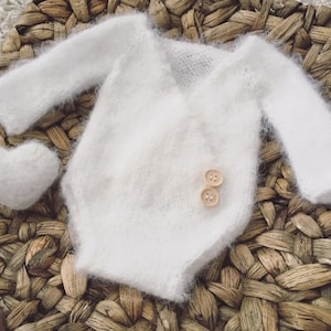Könnte beinhalten: Weißes gestricktes Baby-Outfit mit langen Ärmeln und Wickelverschluss. Das Outfit besteht aus einem weichen, flauschigen Material und hat zwei kleine Holzknöpfe.