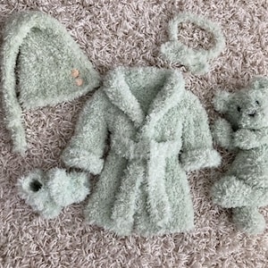 May include: A set of baby clothes in a light green color, including a hooded robe, a hat, booties, and a plush toy. The items are made of a soft, fuzzy material.