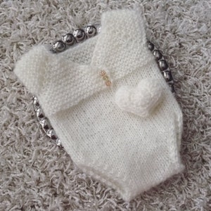 May include: White knitted baby onesie with a heart-shaped applique and pearl embellishments.