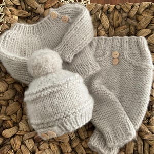 May include: A grey knitted baby outfit with a hat, sweater, and trousers. The outfit has wooden buttons and a pom-pom on the hat.