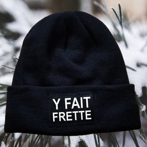 May include: Black beanie with a white patch that says "Y FAIT FRETTE" on it. The beanie is sitting on a snowy surface.
