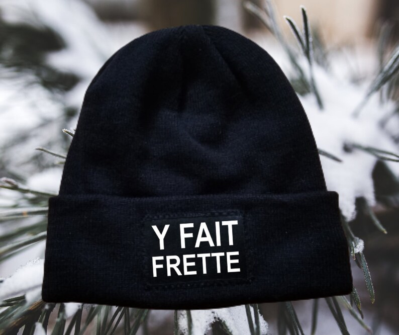 May include: Black beanie with a white patch that says "Y FAIT FRETTE" on it. The beanie is sitting on a snowy surface.