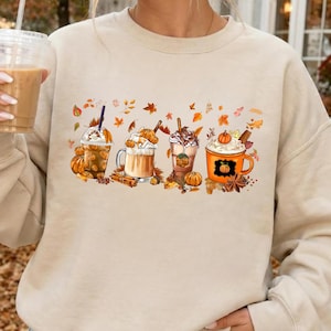 May include: A beige crewneck sweatshirt featuring a graphic print of four different coffee and pumpkin-themed beverages. The design includes various cups, mugs, whipped cream, cinnamon sticks, and scattered autumn leaves in shades of orange and brown.