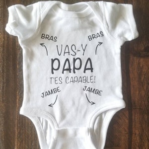 May include: White baby onesie with black text that reads "Vas-y Papa T'es capable!" with arrows pointing to the words "Bras" and "Jambe".