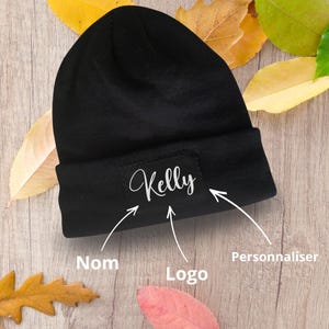 May include: A solid black knit beanie featuring a rectangular patch on the cuff. The patch displays the name Kelly in a white cursive script. Three white arrows point toward the name, the patch, and the cuff with the text Nom, Logo, and Personnaliser.