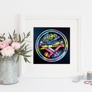 May include: A framed artwork featuring a circular arrangement of colorful crayons. The crayons, labeled "Crayola," are in shades of yellow, blue, pink, and purple. The artwork is set against a black background and framed in white. A bouquet of pink flowers is in a metal pitcher.