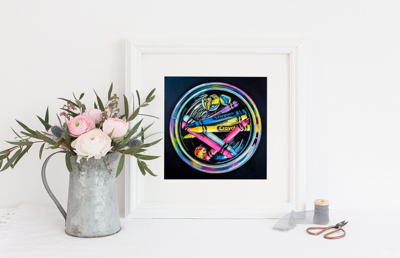 May include: A framed artwork featuring a circular arrangement of colorful crayons. The crayons, labeled "Crayola," are in shades of yellow, blue, pink, and purple. The artwork is set against a black background and framed in white. A bouquet of pink flowers is in a metal pitcher.