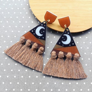 May include: A pair of dangle earrings with a celestial design. The earrings feature a brown and black triangle with a white crescent moon and stars. They have a brown tassel and a brown top.