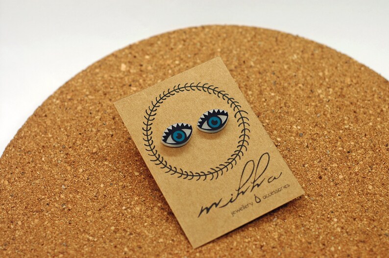 以下が含まれることがあります： 茶色のカードに青い目のスタッドピアスが2つ。カードには「mithra jewellery & accessories」の文字が書かれている。
