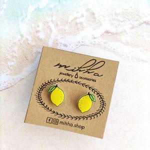 Peut inclure: Boucles d'oreilles clous en forme de citron sur une carte marron. Les boucles d'oreilles sont jaunes avec des feuilles vertes et des contours noirs. La carte porte le texte "mihha jewellery & accessories" et le compte Instagram "mihha.shop".