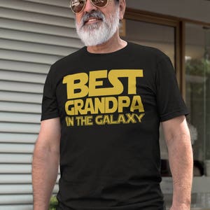 May include: A black t-shirt with the text "BEST GRANDPA IN THE GALAXY" in yellow letters. The person wearing the shirt has a beard and is wearing sunglasses.