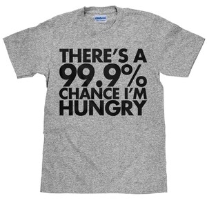 Könnte beinhalten: Ein graues T-Shirt mit dem Text "THERE'S A 99.9% CHANCE I'M HUNGRY" in fetten schwarzen Buchstaben. Das T-Shirt hat kurze Ärmel und besteht aus einem weichen Material. Das Gildan-Logo ist am Kragen sichtbar.