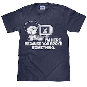 May include: A navy blue t-shirt featuring a white cartoon illustration of a person with glasses holding a computer mouse and an hourglass. The text "I'm here because you broke something." is printed in white below the illustration.