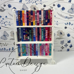 Peut inclure: Une étagère roulante blanche remplie de livres miniatures. Les livres ont des dos colorés et des titres variés. L'étagère a trois étagères et est sur roulettes. Le texte "Custom Design Shop" est au bas de l'image.