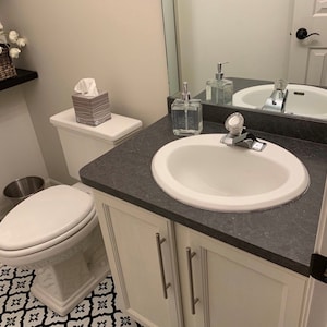 K&ouml;nnte beinhalten: Ein wei&szlig;er Badezimmer-Waschtisch mit einem wei&szlig;en Porzellanwaschbecken und einer wei&szlig;en Toilette. Der Waschtisch hat eine dunkelgraue Arbeitsplatte und zwei wei&szlig;e Schrankt&uuml;ren mit silbernen Griffen. Die Toilette hat einen wei&szlig;en Sitz und einen wei&szlig;en Tank. Auf der Arbeitsplatte steht eine Schachtel mit Taschent&uuml;chern.