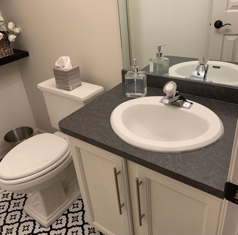 K&ouml;nnte beinhalten: Ein wei&szlig;er Badezimmer-Waschtisch mit einem wei&szlig;en Porzellanwaschbecken und einer wei&szlig;en Toilette. Der Waschtisch hat eine dunkelgraue Arbeitsplatte und zwei wei&szlig;e Schrankt&uuml;ren mit silbernen Griffen. Die Toilette hat einen wei&szlig;en Sitz und einen wei&szlig;en Tank. Auf der Arbeitsplatte steht eine Schachtel mit Taschent&uuml;chern.