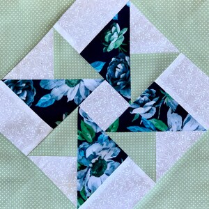 May include: A quilt block with a geometric pattern featuring white, green, and blue floral fabric. The block is made up of triangles and squares.