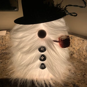 May include: A white furry snowman figurine with a black top hat, a black button nose, and three black buttons for eyes. The snowman is holding a red and white pipe.