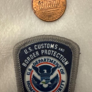 May include: A copper U.S. one-cent coin and a U.S. Customs and Border Protection patch. The coin shows the Lincoln Memorial. The patch is navy blue with white text and a circular emblem featuring an eagle and the words "U.S. Customs and Border Protection".