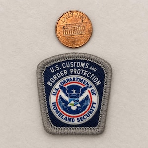 May include: A shield-shaped embroidered patch featuring the U.S. Customs and Border Protection emblem with a central eagle, blue, white, and red details, and a grey border, shown next to a U.S. penny for scale, measuring 0.75 inches in diameter.