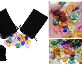 Puede incluir: Un surtido de gemas acrílicas facetadas de colores en varias formas y tamaños, incluyendo corazones, óvalos y redondos. Las gemas se derraman de dos bolsas negras con cordón, con una tercera bolsa visible. Una mano sostiene una selección de las gemas.