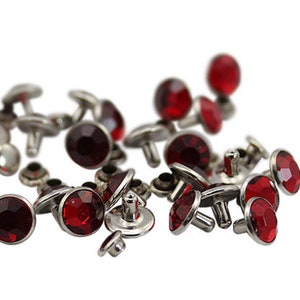 May include: A collection of red rhinestone studs with silver settings. The studs are arranged in a random pattern on a white background.