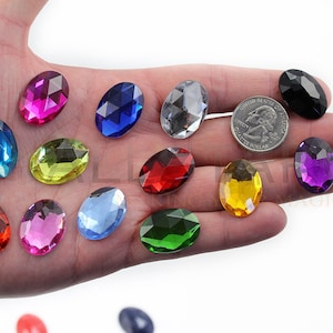 May include: Assortment of oval-shaped, faceted, glass gems in various colors, including red, blue, green, yellow, purple, pink, and clear. The gems are displayed on a hand, with a US quarter for size reference.