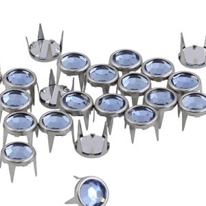May include: A collection of silver-toned metal studs with light blue, faceted, oval-shaped gems. Each stud has three prongs and a flat base. The gems have a slight sparkle, and the studs are scattered against a white background.
