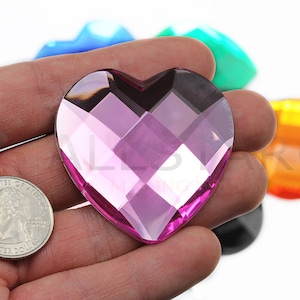 Puede incluir: Una gema rosa en forma de corazón facetado se sostiene en una mano, con una moneda para comparar el tamaño. Otras gemas de colores son visibles en el fondo. La gema tiene una apariencia de vidrio tallado.