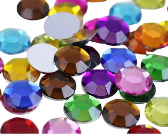 May include: A selection of multi-coloured, faceted rhinestones in various shapes and sizes. The gemstones are in shades of red, blue, green, yellow, purple, and brown. Some are opaque white. The rhinestones are scattered on a white background.