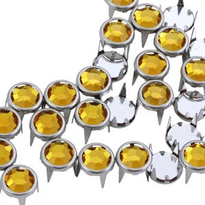 May include: A close-up of a group of amber-colored rhinestones with silver settings. The rhinestones are arranged in a semi-circle shape and have a pointed base.