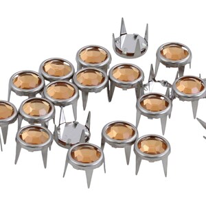May include: A collection of silver-colored metal studs with amber-colored, faceted glass centers. Each stud has a circular top and three pointed prongs. The studs are scattered against a white background.