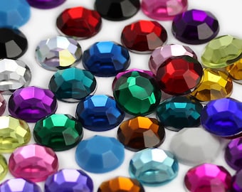 May include: Assortment of colourful, faceted rhinestones in various sizes and shapes. The rhinestones are arranged on a white surface.