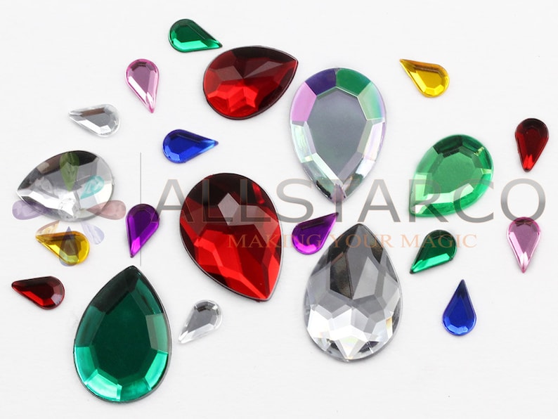 May include: Assortment of teardrop-shaped rhinestones in various colors, including red, green, purple, blue, and clear. The rhinestones vary in size and are scattered across a white surface. The image includes the text "ALLSTARCO MAKING YOUR MAGIC."