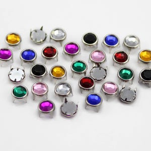 May include: A selection of colourful, round metal studs with faceted, jewel-like centres. The studs are in various colours, including yellow, pink, blue, red, green, and silver, and are arranged on a white surface. Each stud has a silver base with four prongs.