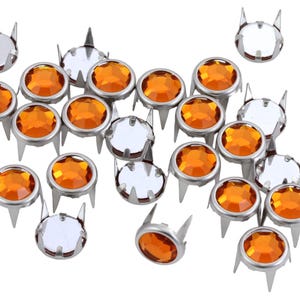 May include: A collection of silver-colored metal studs with orange and clear faceted gems. The studs have a round gem set in a metal bezel with three prongs. The gems are a bright, warm orange color.