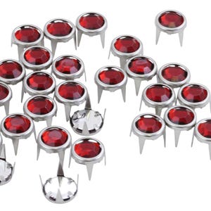 May include: A collection of red rhinestone studs with silver settings. The studs have three prongs on the back for attaching to fabric.