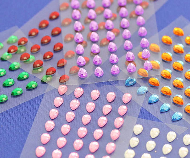 May include: A collection of colorful, teardrop-shaped rhinestones in various colors, including red, green, purple, pink, orange, blue, and white. The rhinestones are arranged in rows on clear adhesive sheets, suitable for crafts and embellishments.