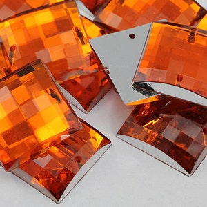 May include: A group of orange, faceted, square-shaped rhinestones with a rounded edge. The rhinestones have two holes on the back for sewing or attaching.