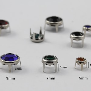 May include: A collection of gemstone rivets in various colors, including red, black, blue, green, and amber. The rivets have a silver-colored metal base with three prongs and are 5mm, 7mm, and 9mm in diameter.